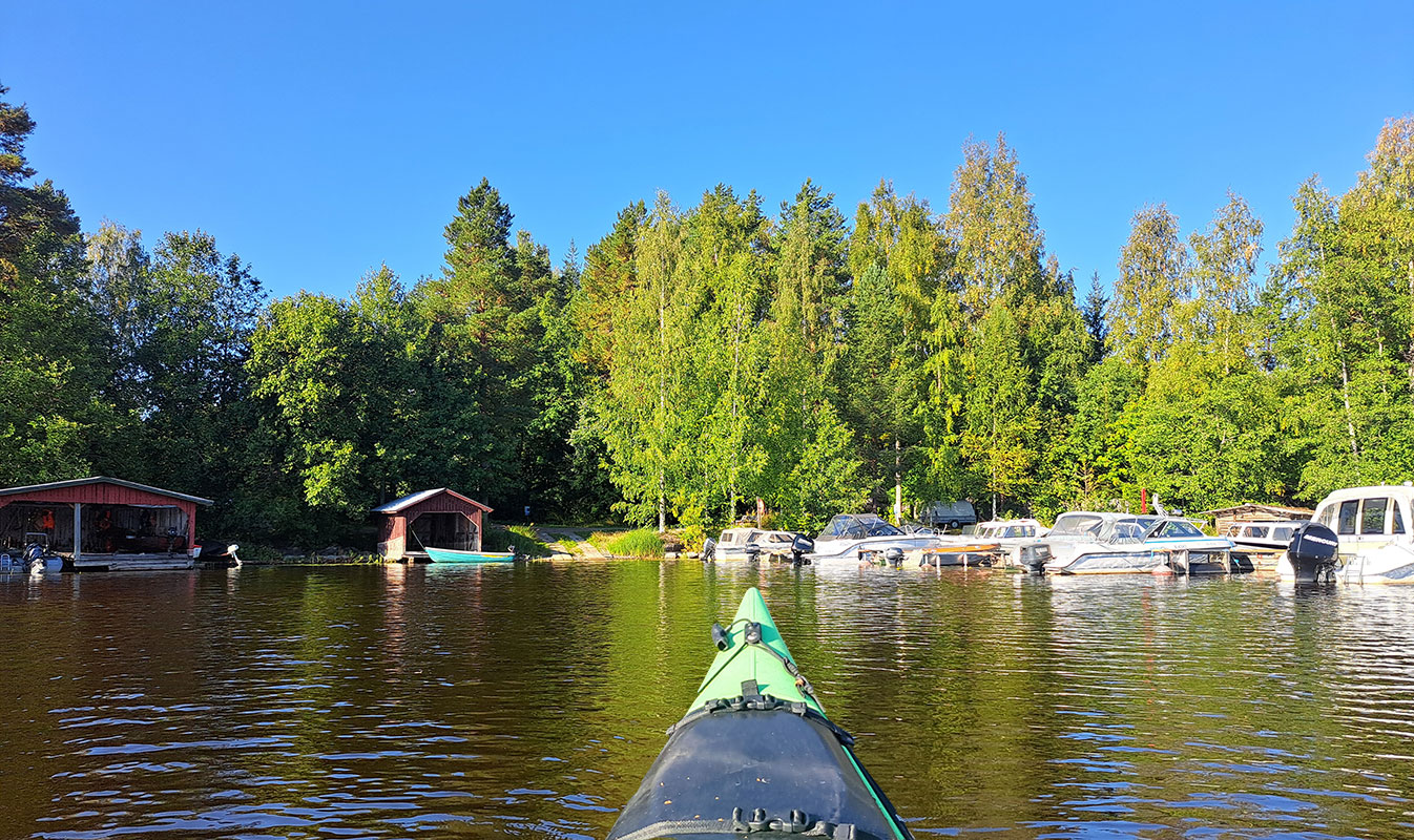 A kayak heads toward Härkinsalmi harbor, with boathouses and boats.