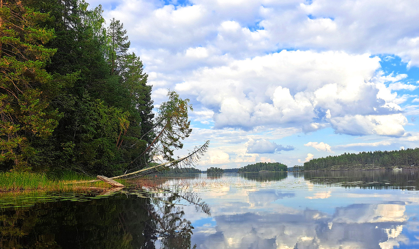 A calm lake view with rocky islets on the horizon. A cloudy sky reflects on the lake’s surface.