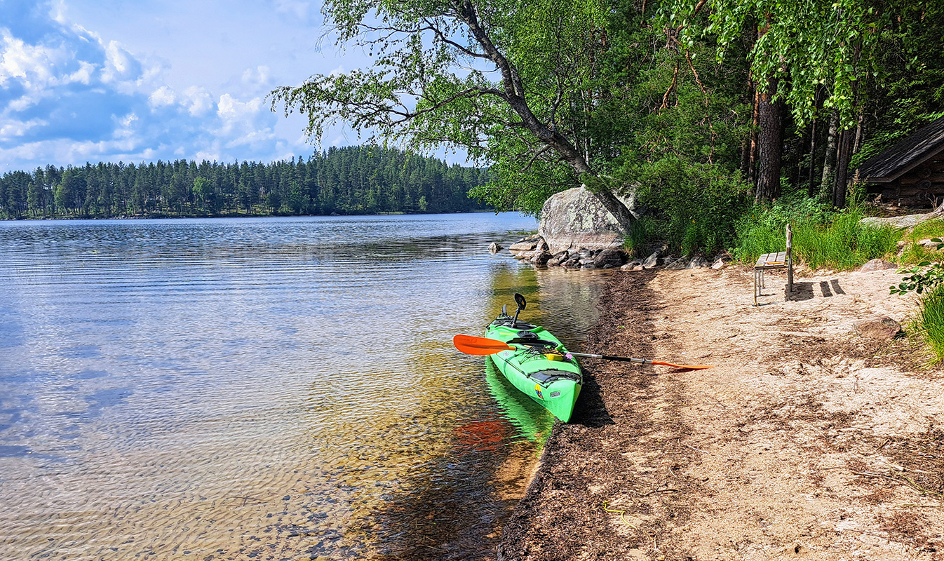 A kayak on a sandy beach, a calm lake, and a forested horizon.