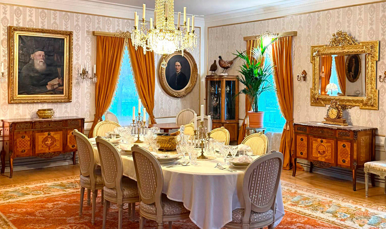 Photo of a manor dining table set for a meal. Paintings on the walls and a large rug under the table.