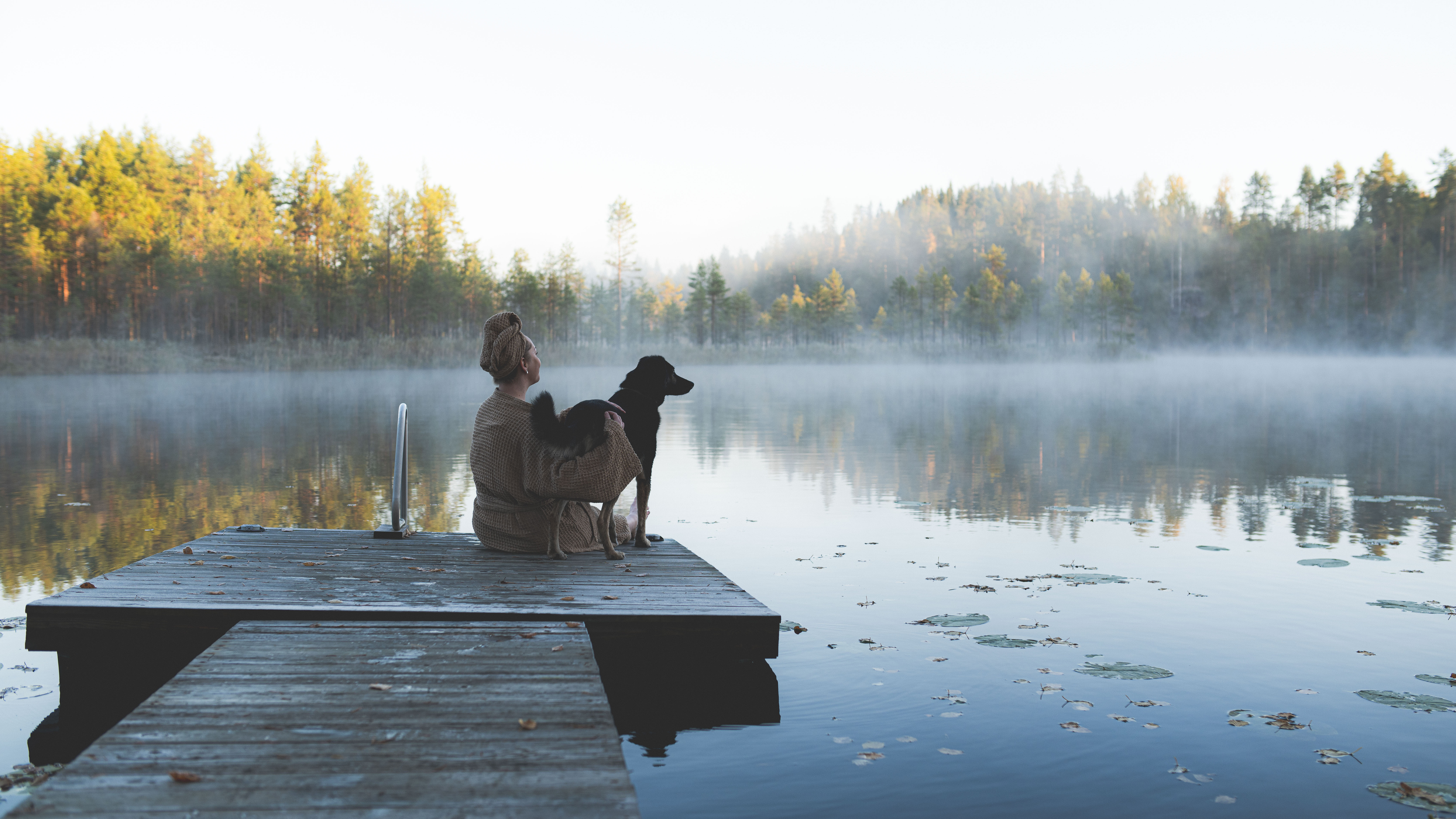 A person sits on a dock with a dog, with a misty lake landscape in the background.